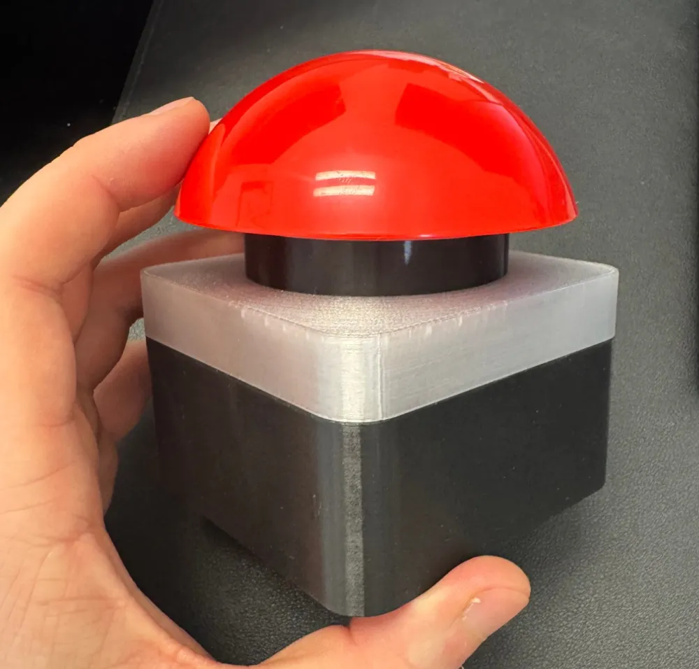 The Guezzer buzzer prototype held in one hand: a glossy red dome on a square black base with a frosted light ring